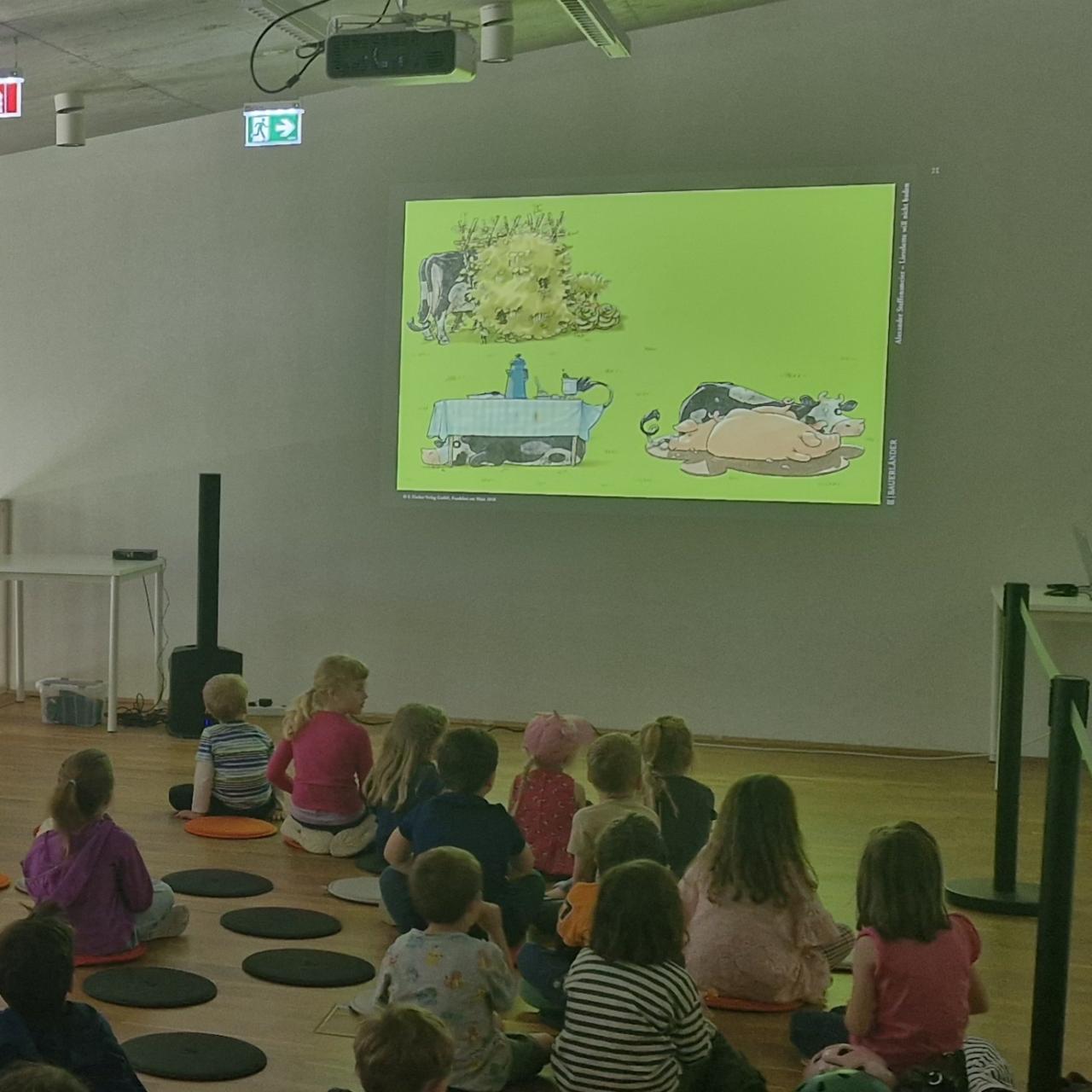 auf der Leinwand sieht man die Kuh Lieselotte mit ihren Tierfreunden, davor sitzen die Kinder und lauschen der Geschichte