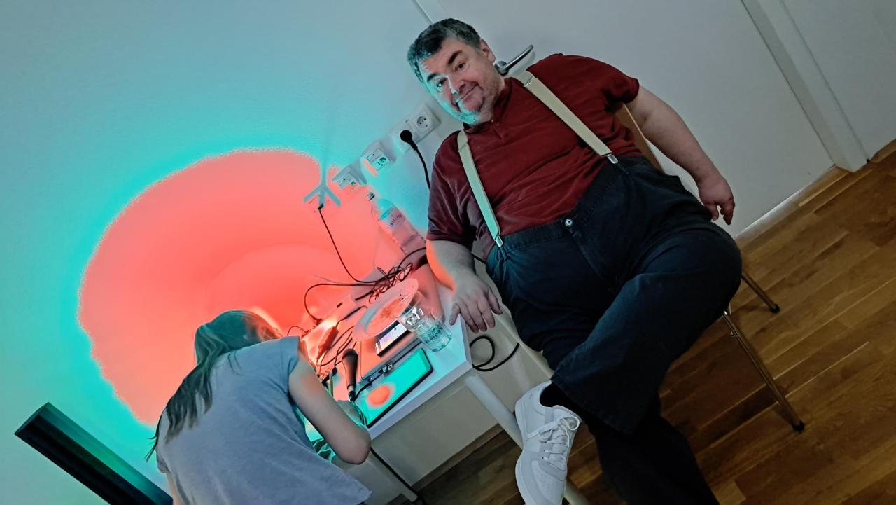 ein Mann sitzt am Computer und ein Kind schreibt einen Liedwunsch auf