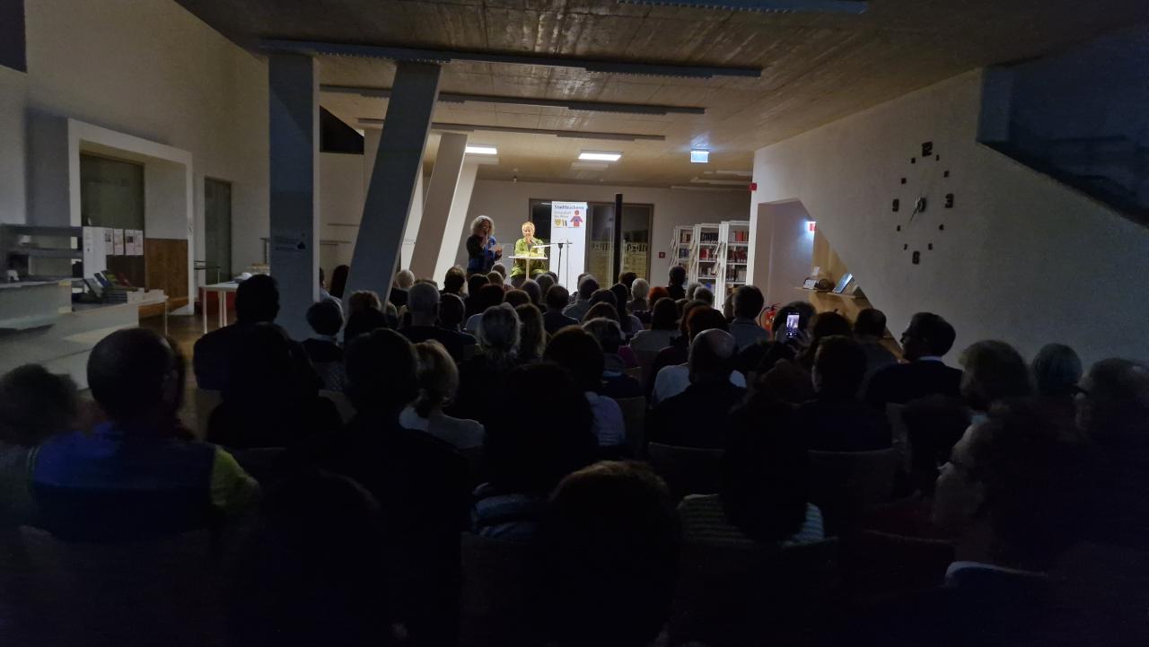 im Vordergrund das Publikum, im Hintergrund sieht man die Büchereileiterin und die Autorin auf der Bühne