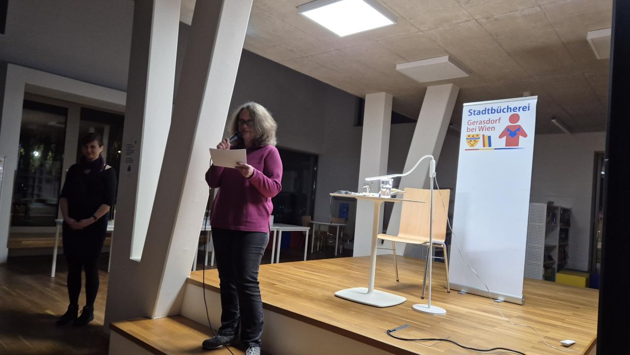 eine Frau steht auf der Bühne, hat einen Zettel und ein Mikrofon in der Hand