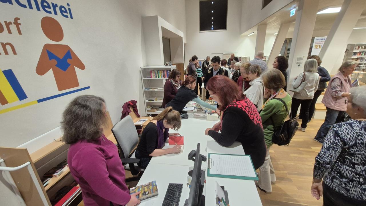 man sieht einen Tisch, es stehen einige Menschen herum, 1 Person sitzt und schreibt