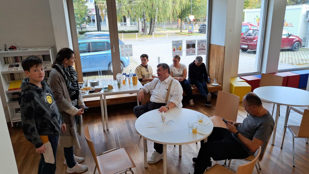 im Hintergrund auf der Fensterbank sitzen 3 Personen, im Vordergrund sitzen 2 Männer um den Tisch und eine Frau und ein Jugendlicher stehen dabei