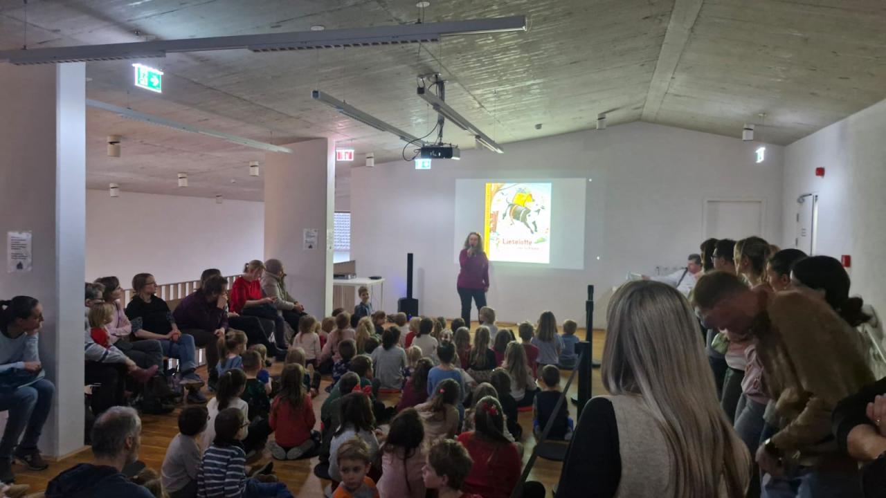 Kinder am Boden und Erwachsene teils auf Sesseln, teils stehend. Vorne steht eine Frau mit Mikrofon.