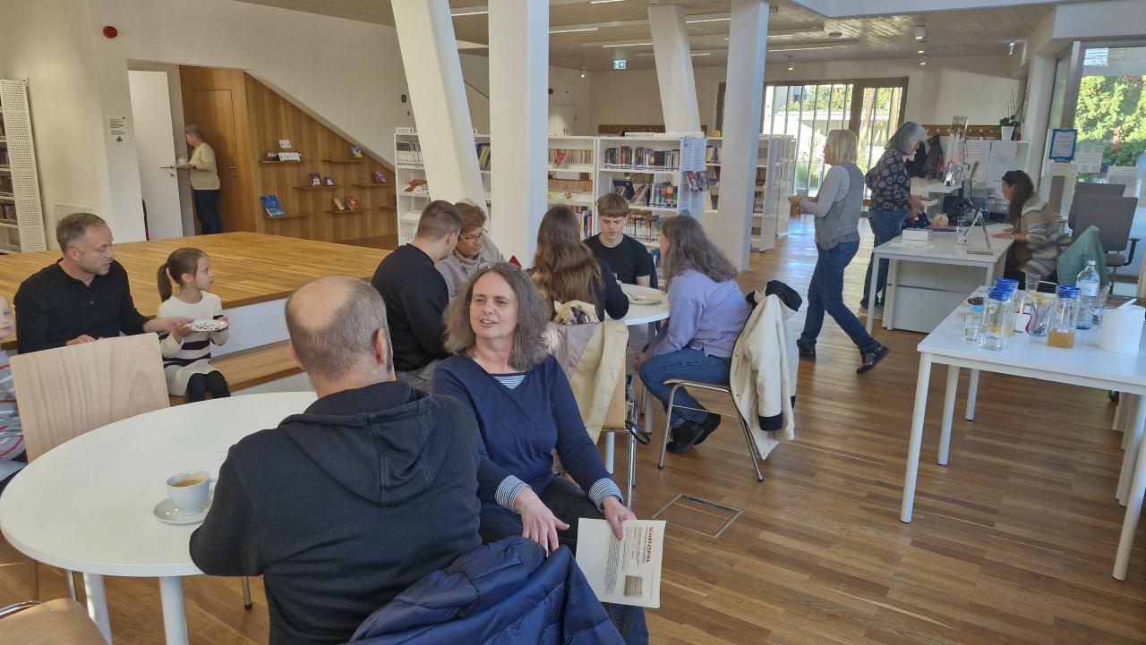 das Foto zeigt Menschen in der Bücherei sitzend, lesend, plaudernd und auch Personen an der Theke beim Verleih