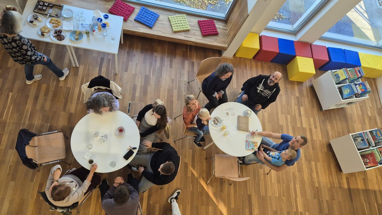 Bild von oben - 2 runde Tische, rundherum sitzen Menschen bei Kaffee und Kuchen und einer netten Plauderei, im Hintergrund das Kuchenbuffet