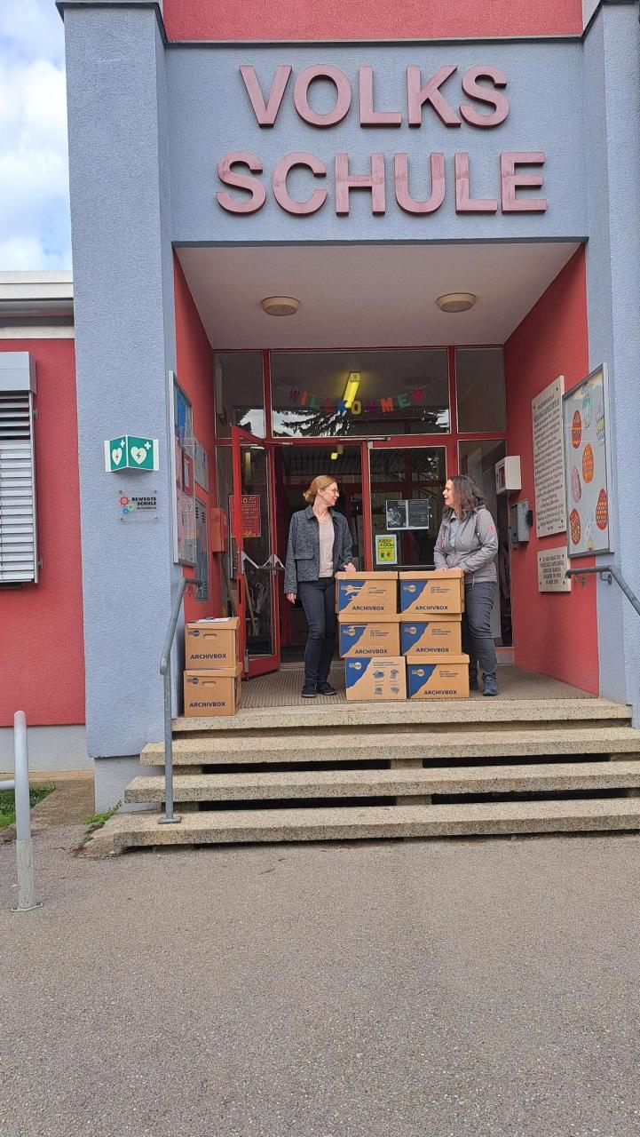 2 Personen stehen mit 6 Bücherkisten vor der Volksschule