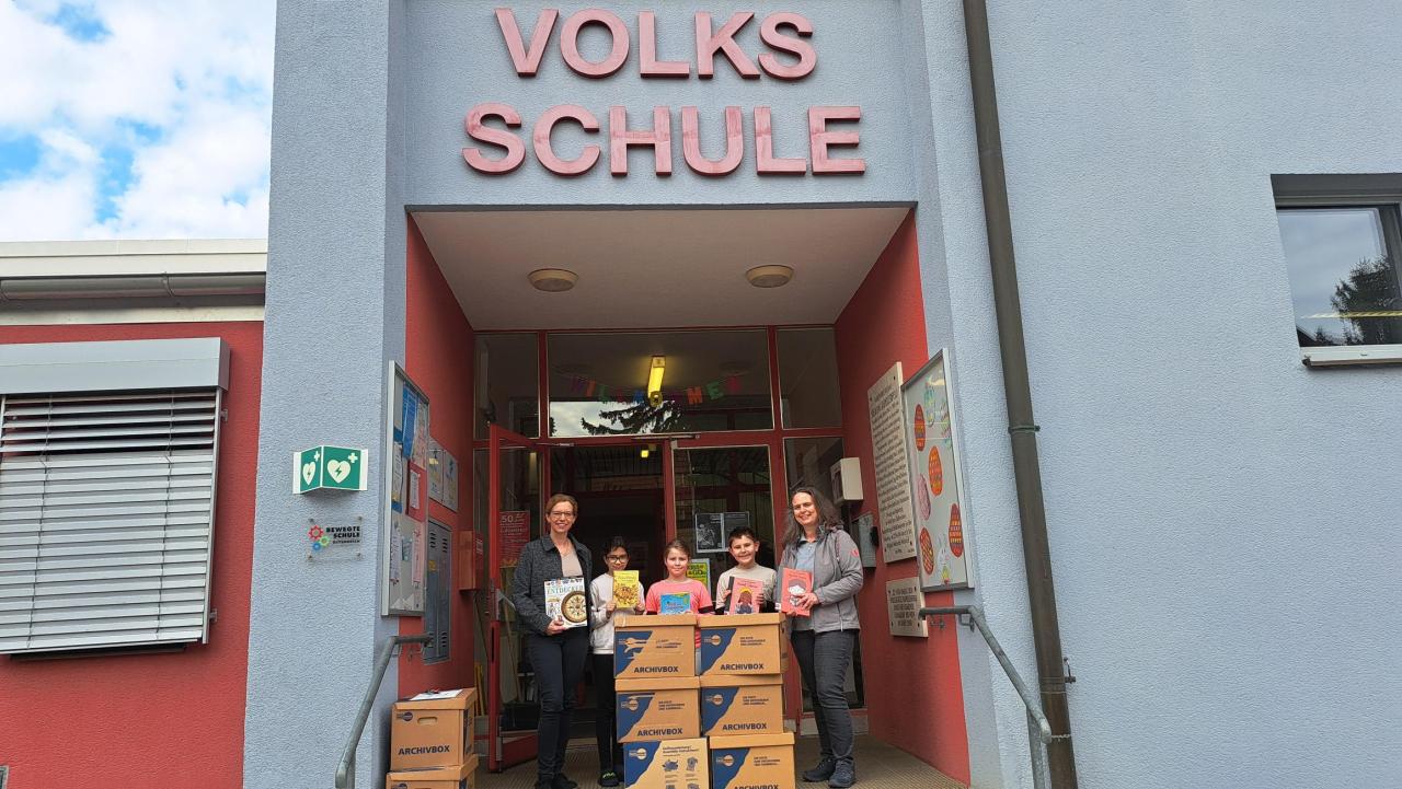 5 Personen haben Bücher in der Hand und stehen vor der Volksschule