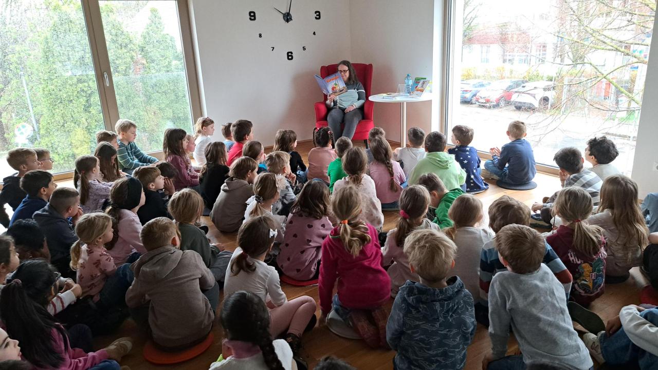 zahlreiche Kinder im Vordergrund, im Hintergrund eine Frau, die ein Buch liest