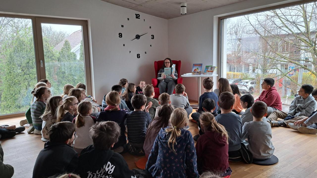 zahlreiche Kinder im Vordergrund, im Hintergrund eine Frau, die ein Buch liest