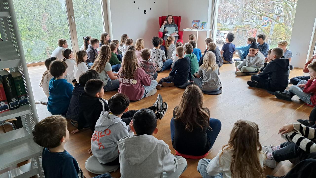 zahlreiche Kinder im Vordergrund, im Hintergrund eine Frau, die ein Buch liest