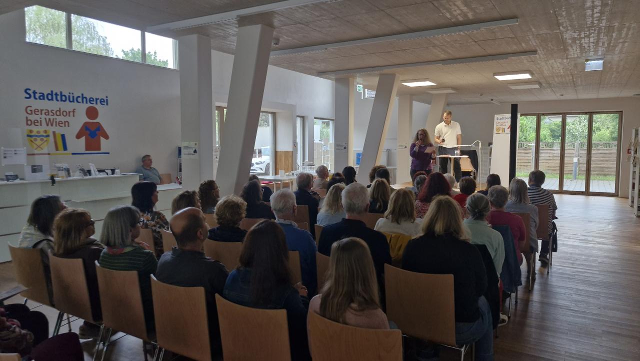 vorne auf der Bühne steht der Autor, davor eine Frau, die das Publikum begrüßt, man sieht das Publikum von hinten
