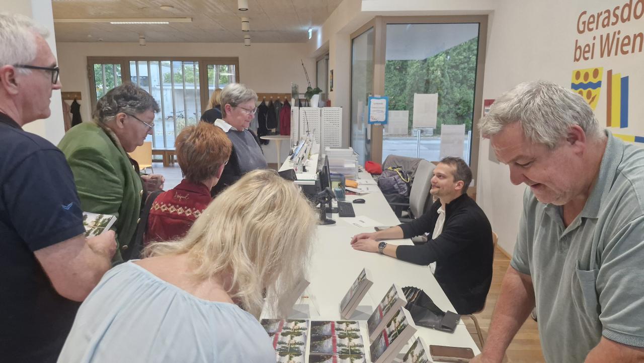 Im Vordergrund sieht man den Büchertisch, wo man das Buch von Lukas Reiff erwerben kann, im Hintergrund sitzt der Autor und ist ins Gespräch mit Fans versunken