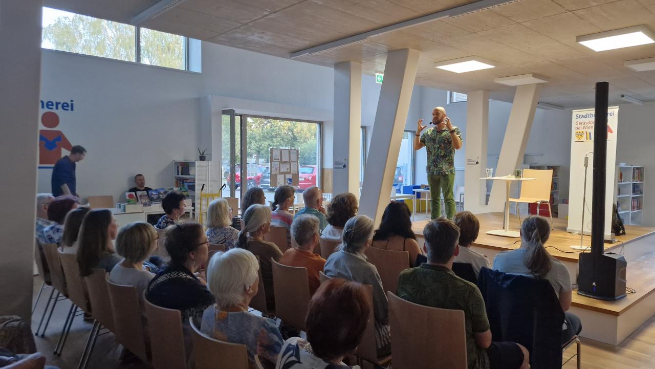 Seitenansicht, der Mann auf der Bühne, das Publikum davor, im hintergrund eine Theke, dahinter 2 Männer, einer sitzt, einer steht