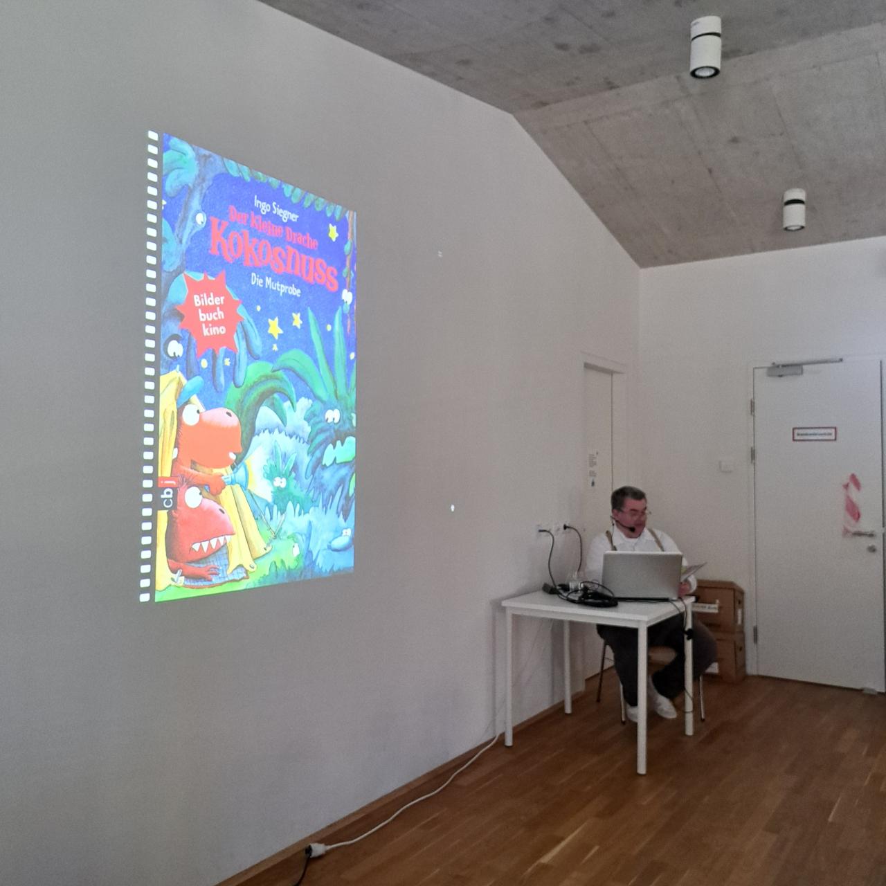 ein Bild mit dem Beamer auf die Wand projiziert, ein Mann sitzt beim Tisch und liest ein Buch